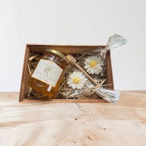 May include: A wooden gift box containing a jar of honey, a wooden honey dipper, and two daisy-shaped soaps. The honey jar has a gold lid and a label that reads "Miele di Acacia Italiano". The soaps are white with yellow centers.