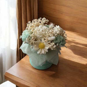 Può includere: Una composizione floreale in una scatola rotonda verde acqua chiaro. Il bouquet presenta fiori bianchi e verde chiaro, tra cui rose e margherite, con piccoli fiori bianchi di riempimento. La composizione è avvolta in carta velina verde acqua chiaro.
