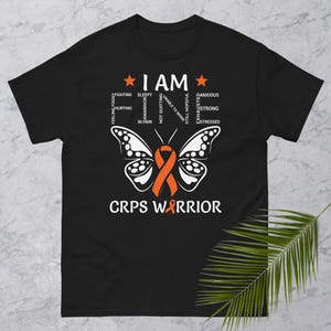 May include: Black t-shirt featuring the text "I AM" above a butterfly design with an orange ribbon. The words "CRPS WARRIOR" are below the butterfly. Additional words include "Fighting", "Sleepy", "Anxious", and "Stressed".