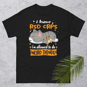 Può includere: Maglietta nera con un gatto dei cartoni animati che indossa un nastro arancione. Il testo sulla maglietta recita "Ho RSD CRPS Sono autorizzato a fare cose strane!"