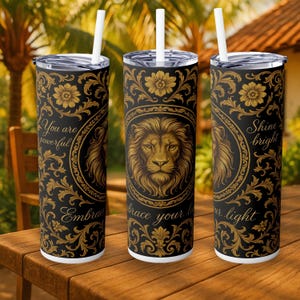 Puede incluir: Tres vasos de acero inoxidable negros con patrones florales dorados e ilustraciones de leones. Cada vaso tiene una pajita blanca y las frases "You are powerful", "Embrace your light" y "Shine bright".