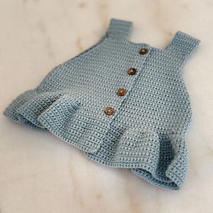 Op de afbeelding: Een lichtblauwe gehaakte babyjurk met een gerimpelde zoom en bruine knopen aan de voorkant.