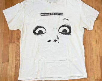 May include: White t-shirt featuring a black and white graphic of a face with wide eyes and the text "AMYL AND THE SNIFFERS" above the eyes. The t-shirt is made of a soft material and has short sleeves.