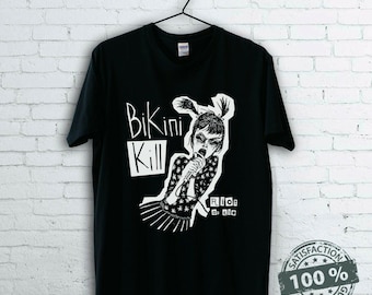 May include: Black t-shirt featuring a white graphic of a woman singing into a microphone, with the words "Bikini Kill" and "Riot Grrrl" in white text. The shirt is hanging on a black hanger.