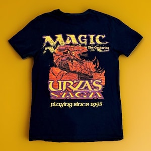May include: Dark blue t-shirt with the text "MAGIC The Gathering" in yellow and a dragon illustration in orange and red. Below the dragon is the text "URZA'S SAGA" in purple and yellow, with "playing since 1993" below.