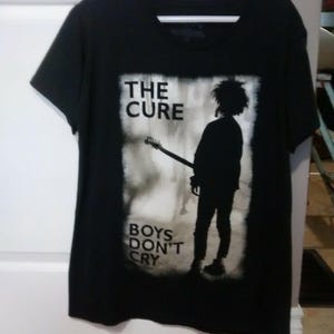 May include: Black t-shirt featuring a graphic of a person playing a guitar, with the words "THE CURE" above and "BOYS DON'T CRY" below. The image is in a rectangular frame on the front of the shirt.