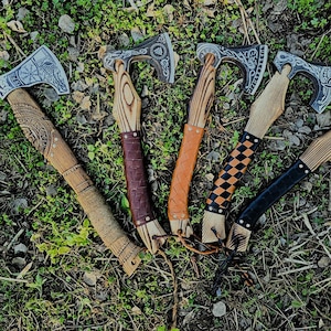 May include: A collection of five Viking axes with wooden handles and intricately designed metal axe heads. The handles feature various leather wraps and decorative carvings. The axes are displayed on a bed of green grass and leaves.