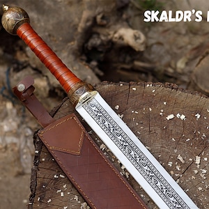 Può includere: Un'immagine dettagliata di una spada con un'impugnatura avvolta in pelle marrone e una lama decorativa. La lama presenta un design intricato. La spada è in un fodero di cuoio marrone. Il testo "SKALDR'S FANG" è visibile.