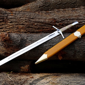 May include: A silver sword with a cross-shaped hilt and a brown handle rests beside its tan leather sheath with gold-colored accents. The sword and sheath are displayed on a background of rough-hewn logs.