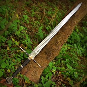 May include: A long, silver sword with a double-edged blade, resting against a tree trunk. The hilt is black with a gold crossguard and a detailed pommel. The sword's blade is approximately 36 inches long.