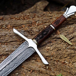 Peut inclure: Gros plan sur une épée avec une lame à motifs et un manche en bois marron. La poignée présente des motifs noirs complexes et est ornée de métal argenté et doré. L'épée repose sur une surface en bois texturée.