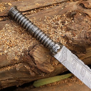 Può includere: Primo piano di un coltello argentato con una lama a motivi e un manico testurizzato. Il coltello è appoggiato su un pezzo di legno con trucioli di legno.