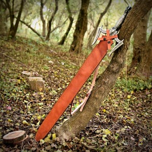 May include: A decorative sword with a silver blade and ornate hilt, sheathed in a brown leather scabbard. The sword is leaning against a tree in a forest setting, with a leather strap attached to the scabbard.