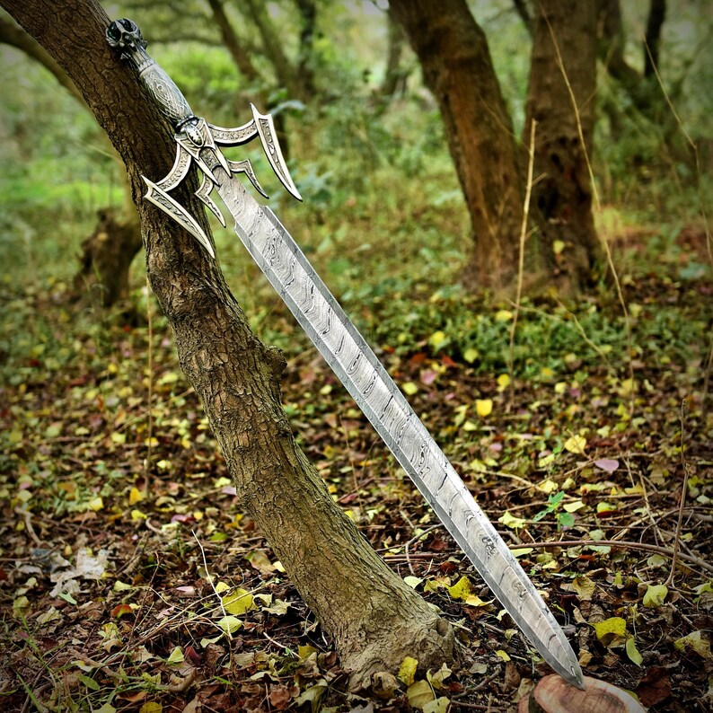 May include: A detailed, silver-colored sword with an ornate handle and a patterned blade, leaning against a tree in a forest setting. The sword features a skull design on the pommel and decorative elements on the guard.