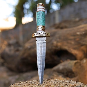 May include: A decorative dagger with a patterned steel blade and a handle featuring turquoise-colored stone, gold-colored metal accents, and a white section. The dagger is displayed against a blurred natural background.