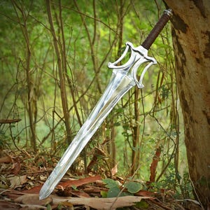Peut inclure: Une épée en métal argenté avec une lame pointue et une garde stylisée. La poignée est enveloppée dans un matériau marron et le pommeau est argenté. L'épée est positionnée en diagonale, avec la pointe reposant sur le sol.
