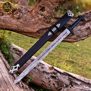 May include: A sword with a patterned Damascus steel blade and a black hilt, accompanied by a matching black scabbard featuring silver-toned metal accents with intricate knotwork designs at the tip and along the length of the sheath.