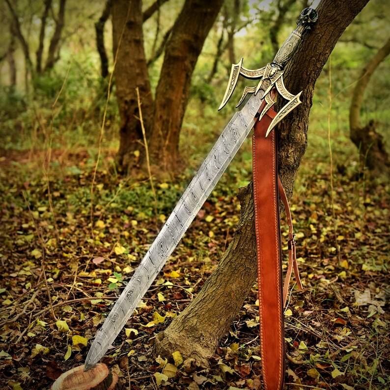 May include: A detailed sword with a patterned blade and ornate gold-colored hilt is displayed against a tree. The sword is paired with a brown leather sheath. The background features a forest setting with fallen leaves.