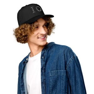 Puede incluir: Una gorra de béisbol negra con las letras IO bordadas en una fuente blanca, texturizada y estampada en el panel frontal. La gorra tiene una corona estructurada, una visera curva y una construcción de tela negra sólida en todo el diseño.