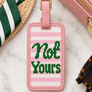 May include: A rectangular luggage tag with a pink and white horizontal striped needlepoint pattern. The text "Not Yours" is stitched in dark green thread across the center. The tag features a pink border and a matching pink strap with a gold buckle.