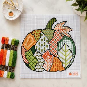 May include: A needlepoint canvas featuring a pumpkin design composed of various geometric and leaf-like patterns in shades of orange, green, and white. The canvas includes the text cara cara CANVAS in the bottom right corner and five embroidery floss skeins.