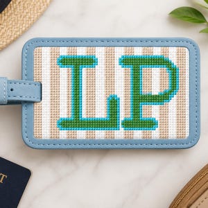 May include: A rectangular luggage tag with a light blue faux leather border and a gold-tone buckle strap. The centre features a beige and white vertical striped background with the letters LP embroidered in green with a bright blue outline.