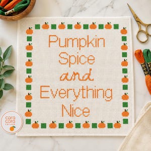 May include: A square cross-stitch embroidery on white fabric featuring the orange text Pumpkin Spice and Everything Nice. The design is framed by a border of alternating small orange pumpkins and green squares. A circular logo reads cara cara canvas.