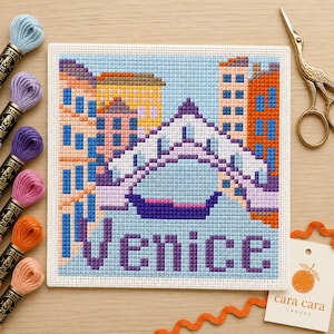 May include: A square needlepoint canvas featuring a colorful cross-stitch design of a Venetian bridge over water with surrounding buildings. The word Venice is stitched in purple at the bottom. Includes embroidery floss skeins and gold scissors.