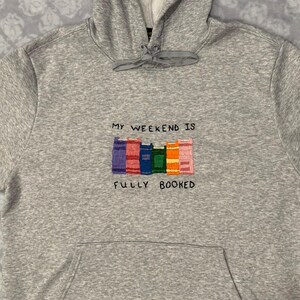 May include: Gray hooded sweatshirt with the text "My weekend is fully booked" embroidered on the front. The text is surrounded by a row of colorful books.