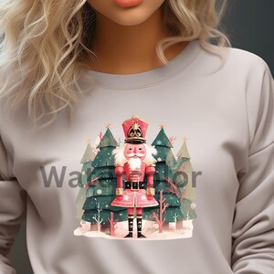 May include: A light gray sweatshirt featuring a colorful graphic of a nutcracker soldier standing in front of Christmas trees. The text "Waterfor" is printed on the sweatshirt. The sweatshirt is a crew neck style.