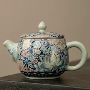 Puede incluir: Tetera de cerámica con diseño floral. La tetera presenta un fondo azul con varias flores en tonos rosa, blanco y azul. El asa y el pico son de color verde claro. La tapa tiene un pequeño pomo en la parte superior. Aproximadamente 15 cm de altura.