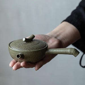 Puede incluir: Pequeña tetera de cerámica verde oliva con tapa y asa larga en ángulo. La tetera tiene una superficie texturizada y un pequeño pico. La tapa presenta un asa curvada. La tetera está sostenida en la mano de una persona.