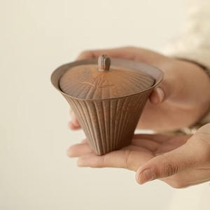 May include: A brown ceramic tea cup with a lid. The cup has a ribbed, cone-like shape and a small knob on the lid. The cup is held in a person's hands. The cup is a warm brown colour.