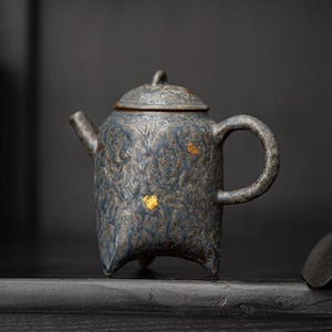 以下が含まれることがあります： テクスチャ加工された表面と蓋が付いた、小さな青いセラミックティーポット。 ティーポットは丸みを帯びたボディ、湾曲したハンドル、注ぎ口を備えています。 繊細な模様と小さな金色のアクセントで装飾されています。 ティーポットは3つの小さな足の上にあります。