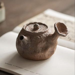 May include: A small, handcrafted brown ceramic teapot with a textured surface. The teapot features a rounded body, a lid with a small knob, and a wide, flared spout. It rests on an open book with Japanese text.