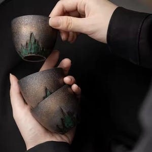 Puede incluir: Tres pequeñas tazas de té de cerámica moteada. Las tazas son de color marrón oscuro con un diseño de montaña verde en la base. Una taza es sostenida por una mano, mientras que las otras dos están apiladas en otra mano.