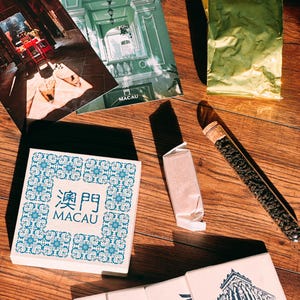 Puede incluir: Una colección de artículos decorativos con temática de Macao. Los artículos incluyen una pequeña caja con un patrón de azulejos azules y blancos, una postal con una foto de un edificio, una postal con el texto "Hi Jill, I'm currently in Macau!" y una pequeña bolsa de té.