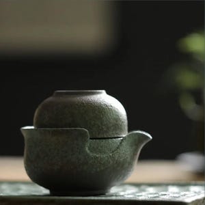 Op de afbeelding: Een donkergroene keramische theeset met een theepot en een kopje. De theepot heeft een tuit en een handvat. Het kopje is rond en heeft geen handvat.