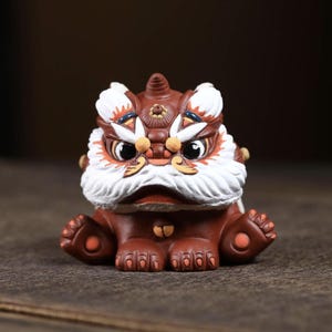 May include: A detailed, brown ceramic lion figurine with white and orange accents. The lion has a white beard, orange eyes, and a detailed mane. The figurine is sitting on a wooden surface.
