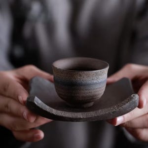 Peut inclure: Une petite tasse à thé en céramique rustique avec un extérieur brun foncé et un intérieur plus clair, posée sur une soucoupe carrée assortie. La tasse et la soucoupe ont un aspect texturé et fait à la main, suggérant des matériaux naturels et un savoir-faire traditionnel.