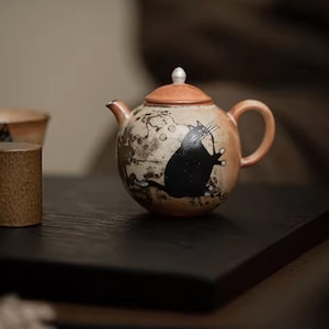 May include: A ceramic teapot featuring a black cat design. The teapot has a rounded shape with a light brown base and a dark brown lid. The cat is illustrated in a playful pose. The teapot includes a handle and a spout.