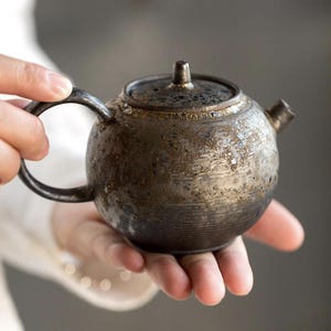 Puede incluir: Una tetera pequeña, redonda y de color oscuro con tapa y asa. La tetera tiene una superficie texturizada y un pico. El asa tiene forma curva y circular. La tetera está sostenida en las manos de una persona.