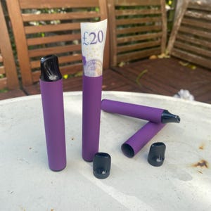 Puede incluir: Varios vape pens desechables morados con boquillas y tapas negras se exhiben sobre una superficie blanca. Un billete de 20 £ está enrollado dentro de uno de los bolígrafos. Los bolígrafos son cilíndricos y tienen un acabado mate.