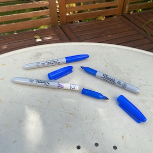 以下が含まれることがあります： 青いシャープのファインポイントの永久マーカーが4本、そのうちの1本には2ポンド札が入っています。マーカーは白い表面の上にあります。