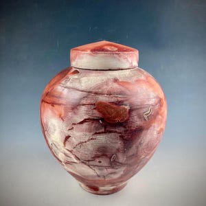 May include: A handcrafted, lidded urn with a rounded body and a triangular top. The urn features a blend of red, white, and brown hues, with a marbled effect. The ceramic vessel is a decorative piece.