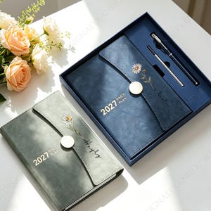 May include: A dark blue 2027 daily planner in a matching gift box with a silver pen and a refill, alongside a sage green 2027 daily planner. Both planners feature a magnetic closure, a floral illustration, and a name printed on the front cover.