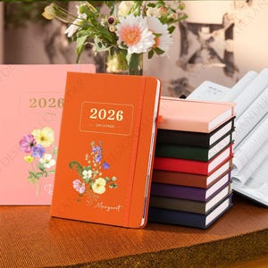 May include: An orange 2026 day-a-page planner with floral design and the name "Margaret" printed on the cover. A stack of colourful notebooks and a pink planner are also visible. A vase of flowers is in the background.