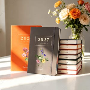 May include: Two hardcover planners with elastic bands, one orange and one dark grey, both labelled 2027 Day A Page with floral illustrations and names. A stack of similar planners sits beside them, with a glass vase of colourful roses in the background.