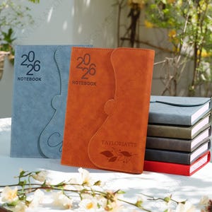 May include: A selection of notebooks in shades of blue, orange, and grey. Each notebook is embossed with the year "2026" and the word "NOTEBOOK". The orange notebook features the name "TAYLORJAYTS" and a leaf design.