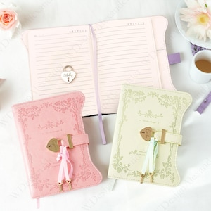 Könnte beinhalten: Rosa und cremefarbene Journale mit floralen Designs und herzförmigen Schlössern. Ein Journal ist geöffnet und zeigt linierte Seiten. Die Journale haben goldfarbene Verschlüsse und dekorative Bänder. Eine Teetasse und Blumen sind im Hintergrund.